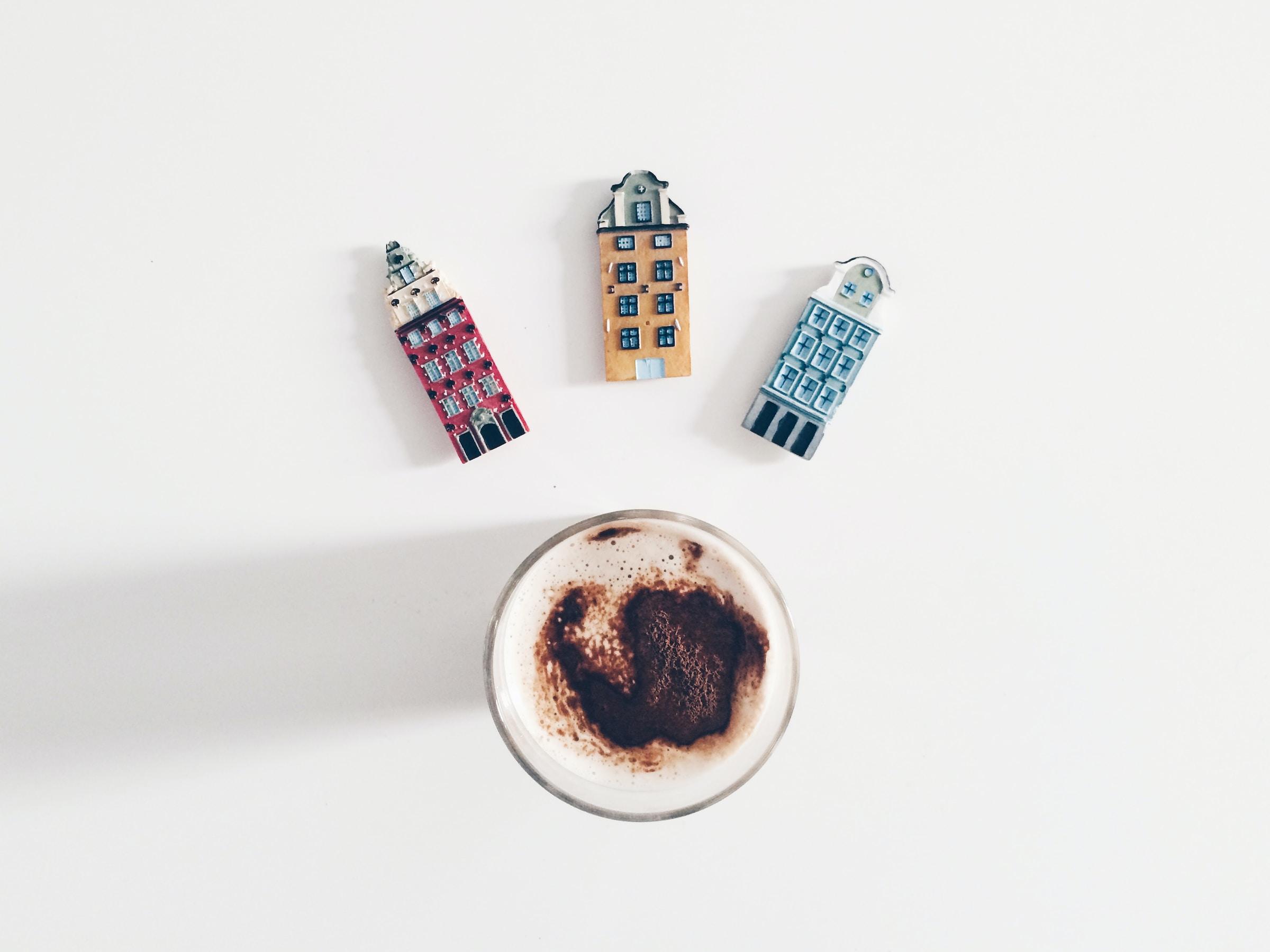 Bovenaanzicht van een kop koffie op een witte tafel. Bij de kop liggen leuke miniatuur kunstwerken van oude huisjes.